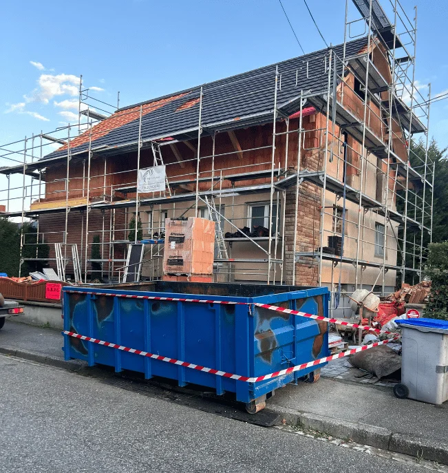 Benne bleue posée devant une maison alsacienne en cours de rénovation, échafaudage et rubalise de chantier.
