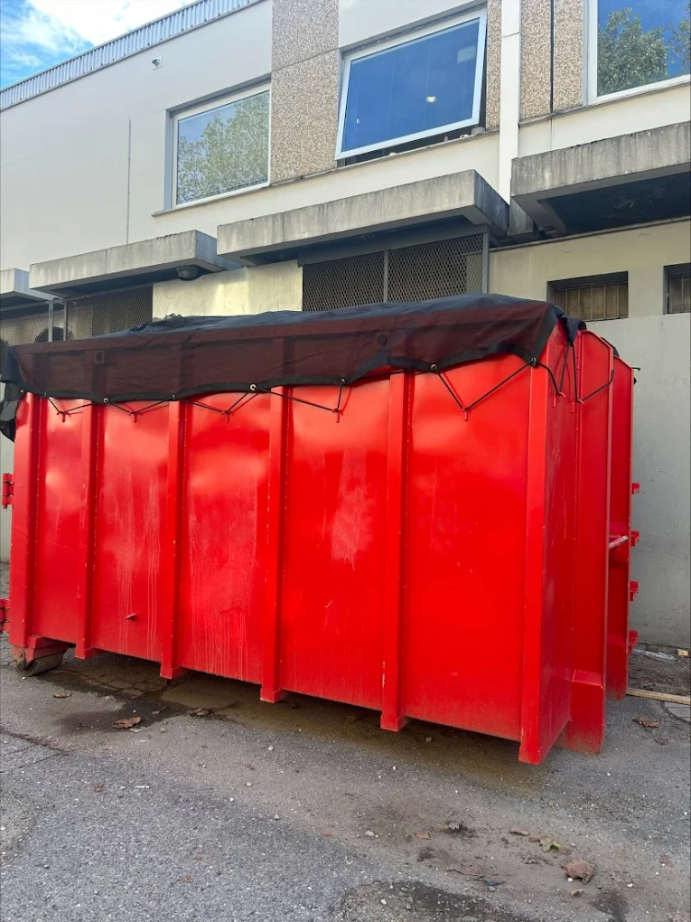 Benne rouge fermée par une bâche noire, posée en pied d'immeuble.